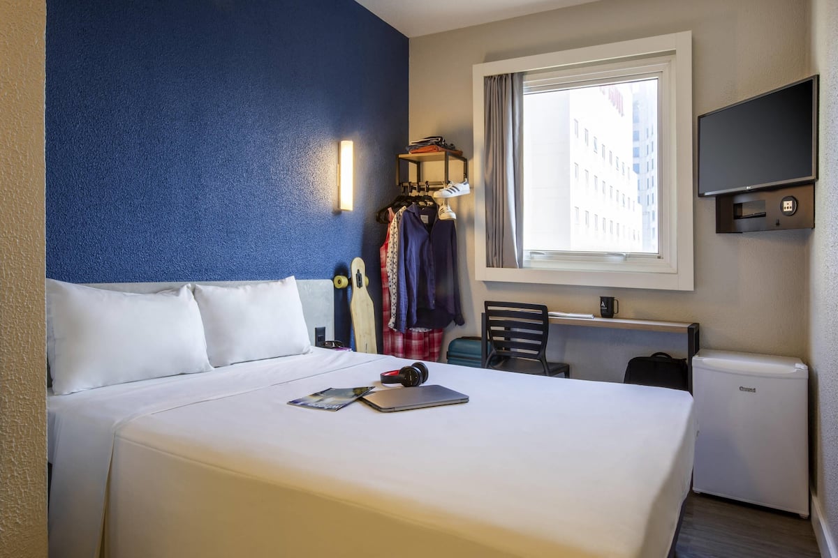 Habitación de hotel con cama, escritorio, silla, televisor y ventana con vista a edificios.