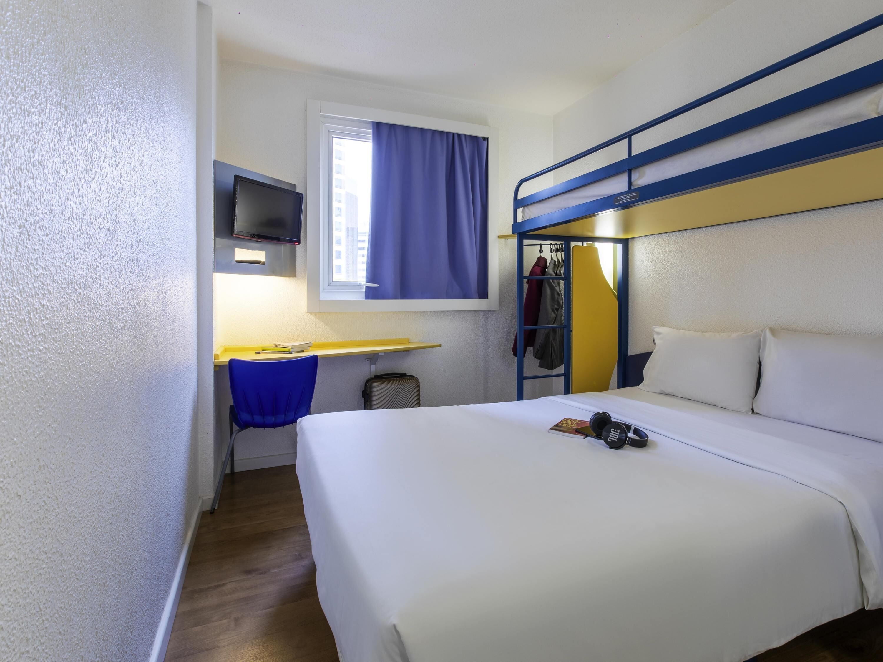 A hotel room with a bunk bed, a desk with a chair, a television, and a window with curtains.