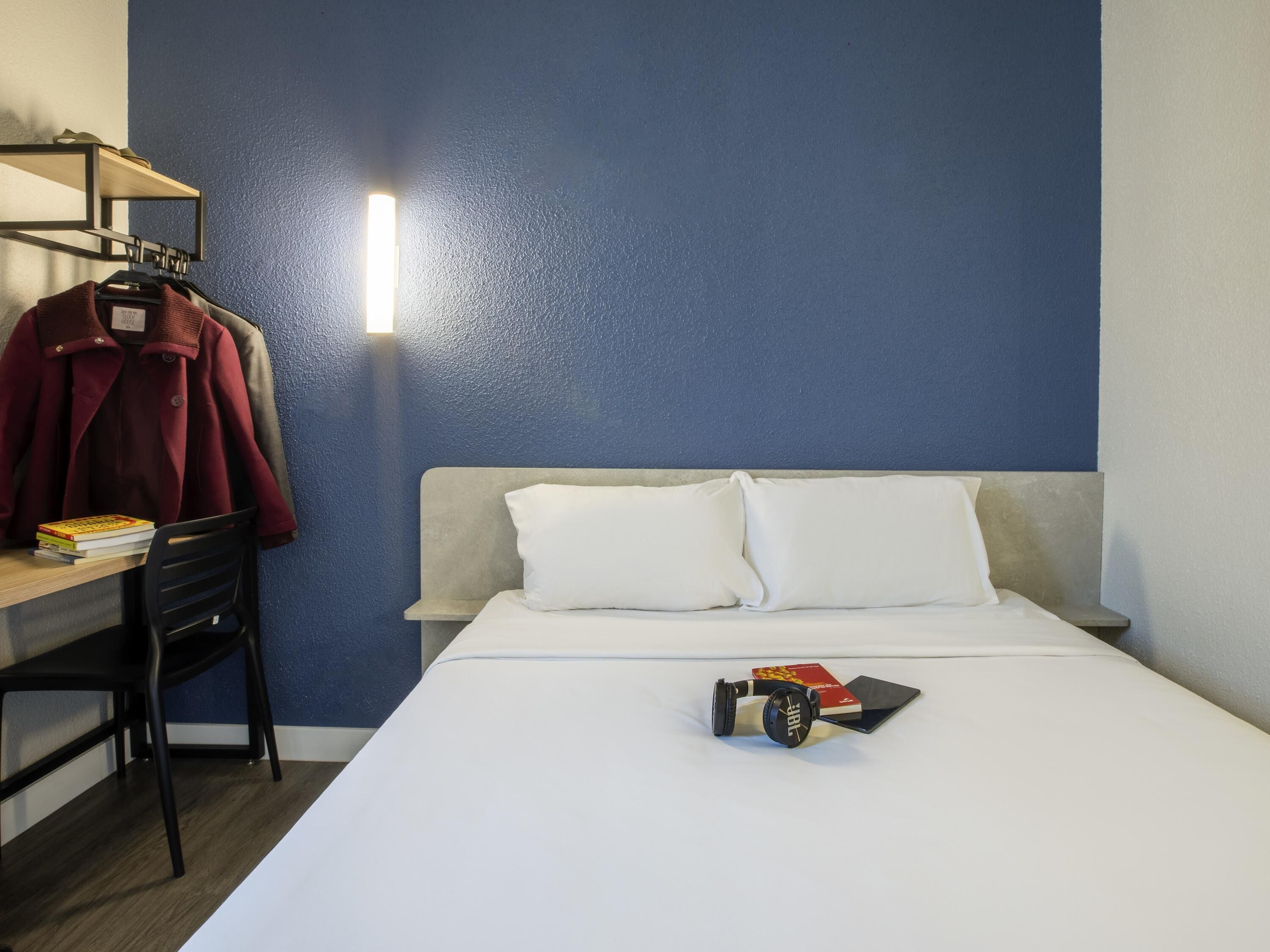 A hotel room with a bed, a desk with a chair, a coat rack with a jacket, and a lamp.