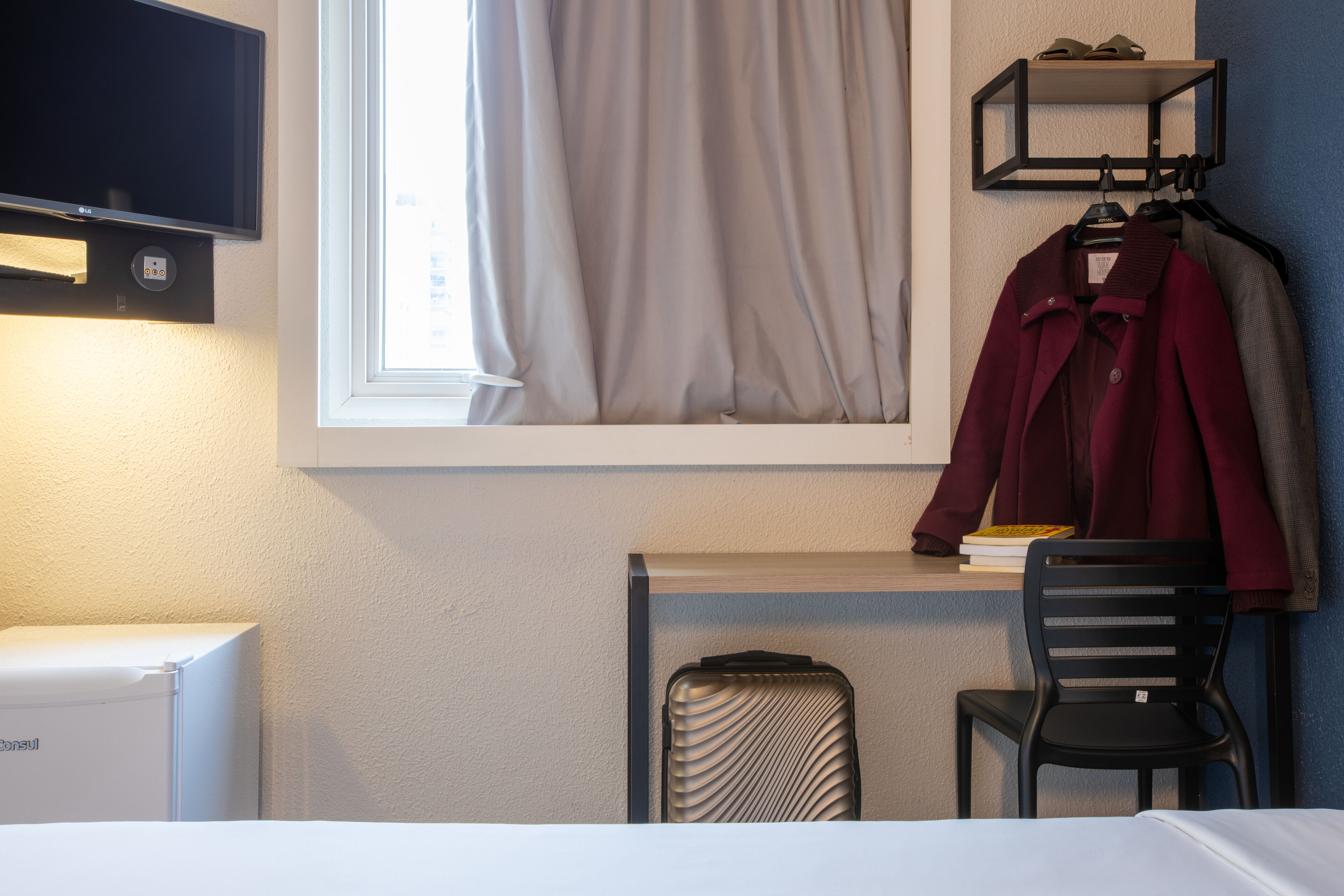 Ein Hotelzimmer mit einem Bett, einem Schreibtisch mit Stuhl, einem an der Wand befestigten Fernseher, einem Fenster mit Vorhängen, einem Kleiderständer mit Kleidung und einem Koffer.