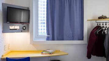 Quarto de hotel com cama, escrivaninha com cadeira, televisão na parede, janela com cortinas e cabideiro com roupas.