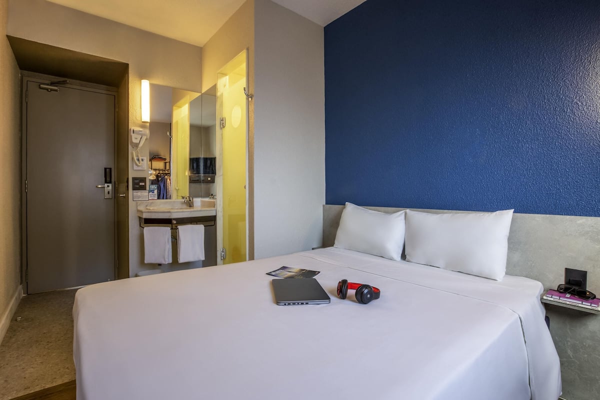 Habitación de hotel con cama, mesita de noche con un libro y auriculares, espejo, lavabo y puerta.