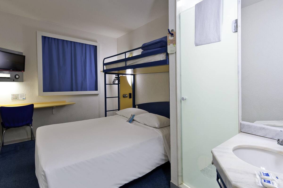 A hotel room with a bunk bed, a desk with a chair, a TV, and a bathroom with a sink and mirror.