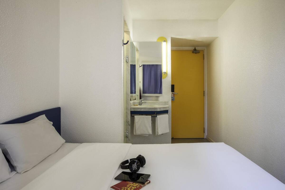 A hotel room with a bed, a bathroom visible through an open door, and a small table with a book and headphones.