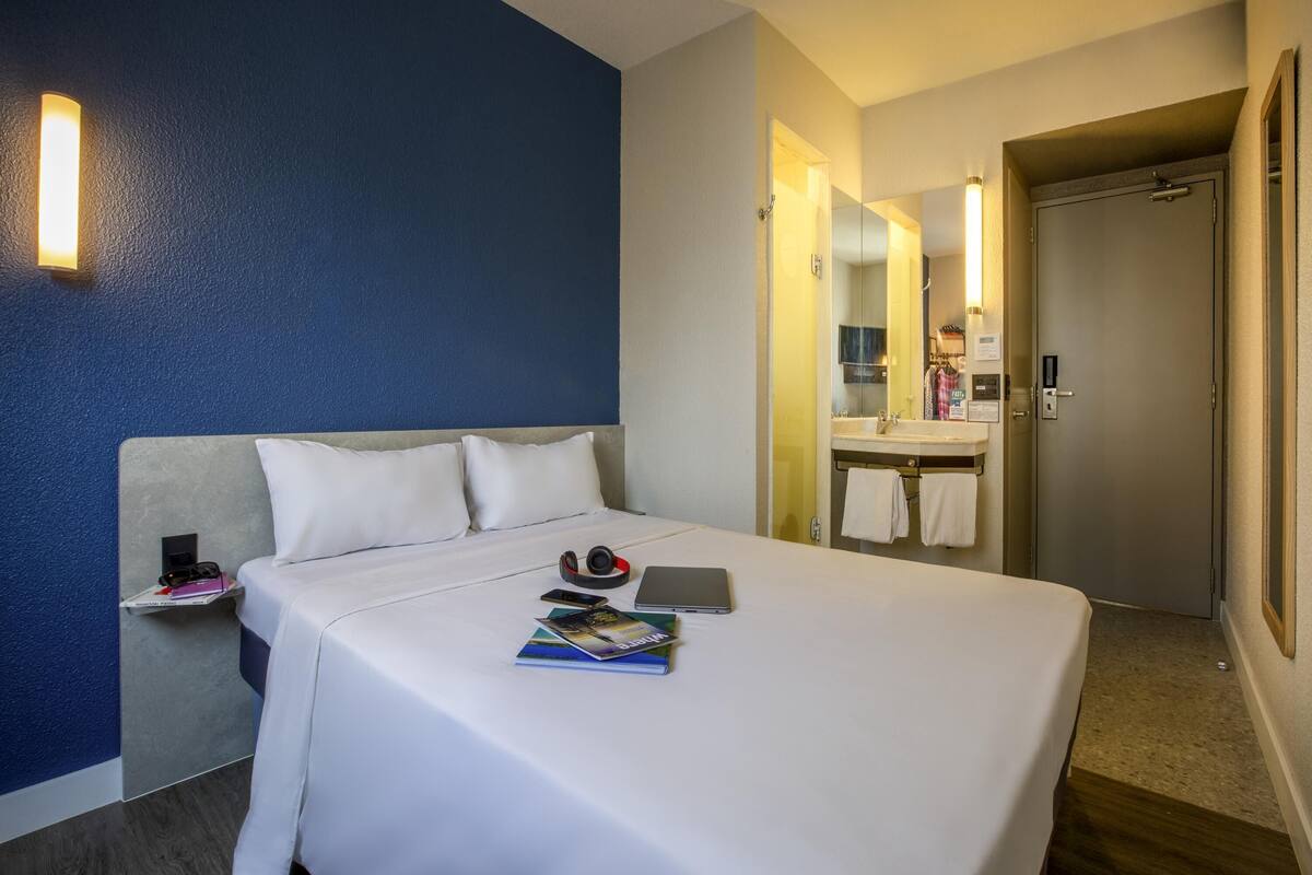 A hotel room with a large bed, a bedside table with a lamp, a mirror, and a bathroom with a sink and towel rack.