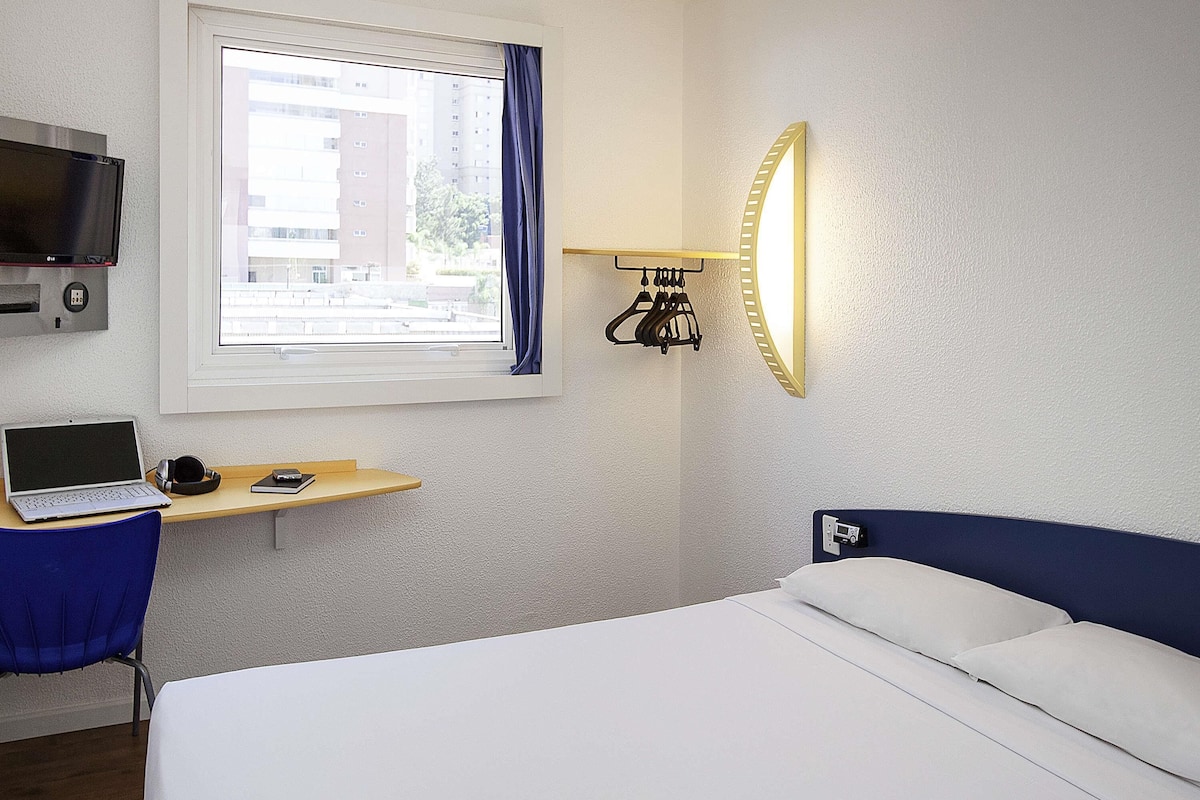 Habitación de hotel con cama, escritorio, laptop, televisor y ventana con cortinas.