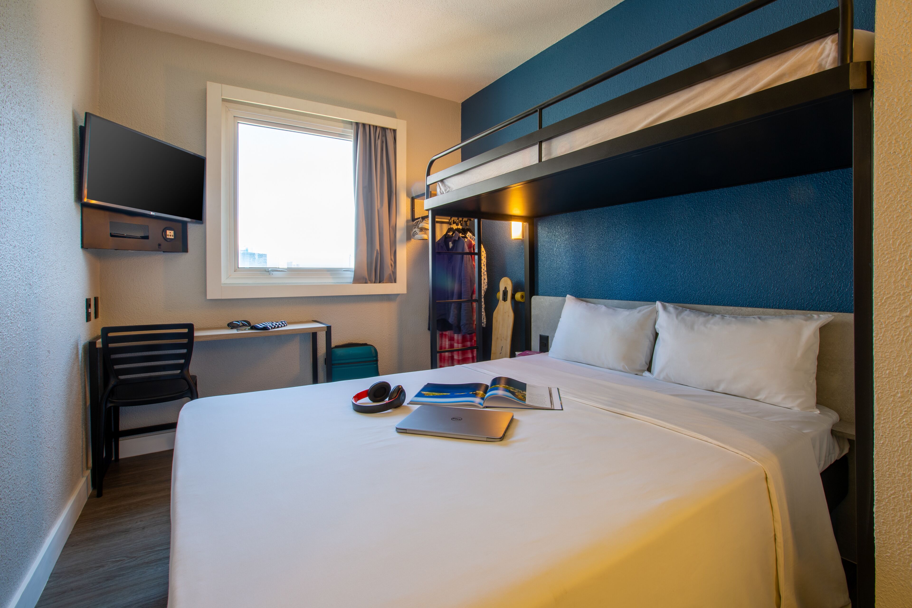 A hotel room with a bunk bed, a desk with a chair, a TV mounted on the wall, and a window with curtains.