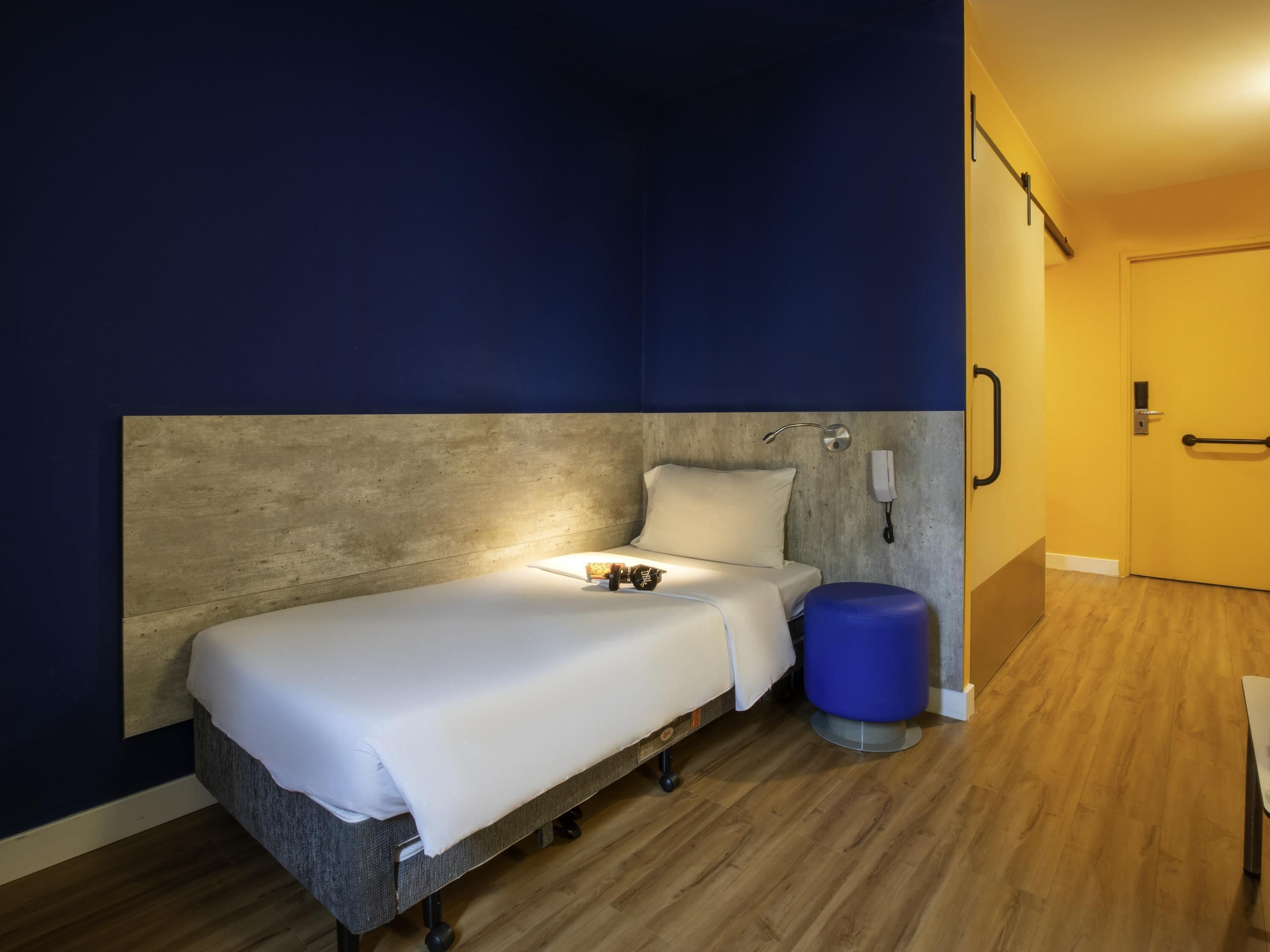 Una cama individual con cabecera, una mesita de noche con lámpara, un teléfono en la pared y una puerta con manija.