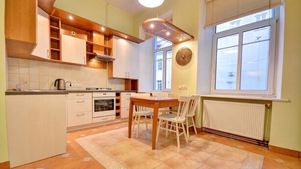 A kitchen with wooden cabinets, a dining table with chairs, a window with blinds, and a radiator. - Great Apartment near Town Hall (Vilnius)