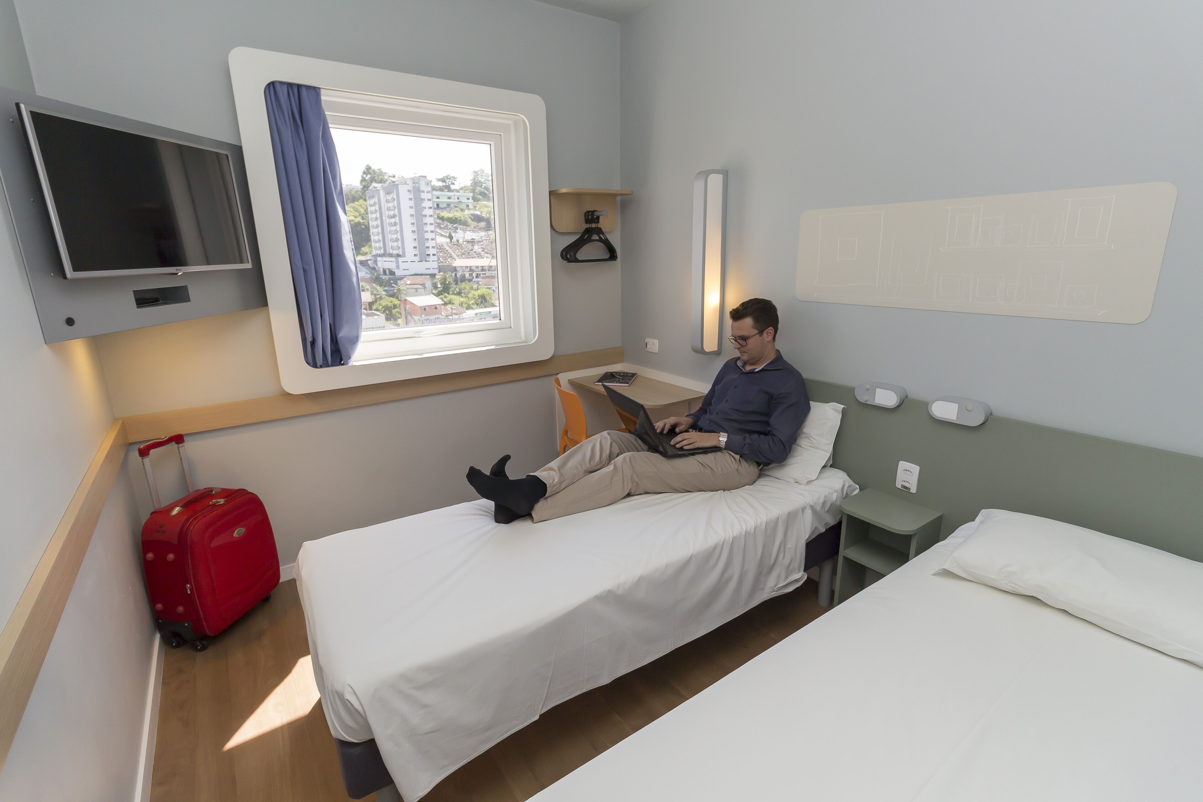 Een hotelkamer met een bed, een bureau, een televisie, een raam met gordijnen en een rode koffer.