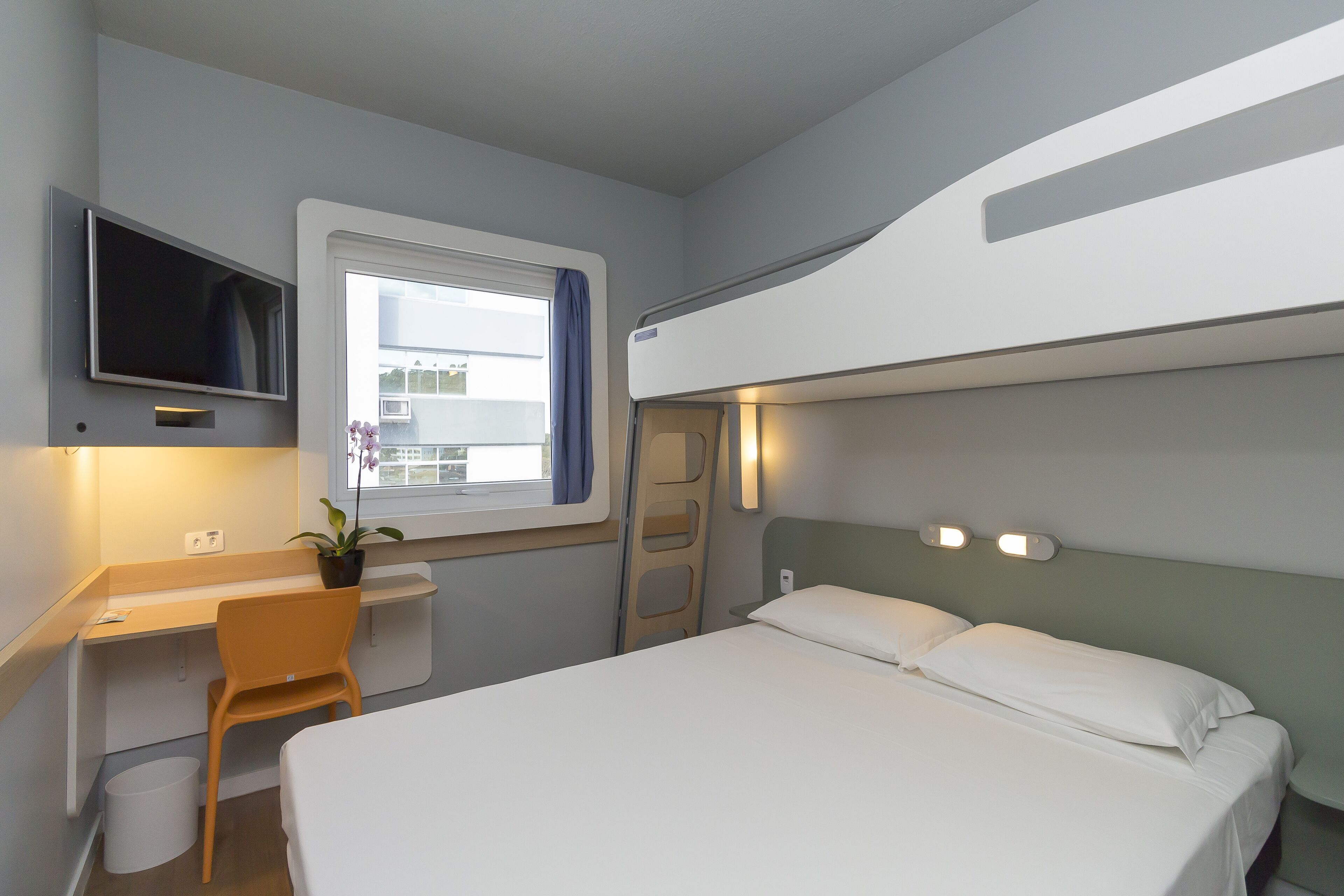 Ein kleines Hotelzimmer mit Bett, Schreibtisch, Stuhl, Fernseher und einem Fenster mit Vorhängen.