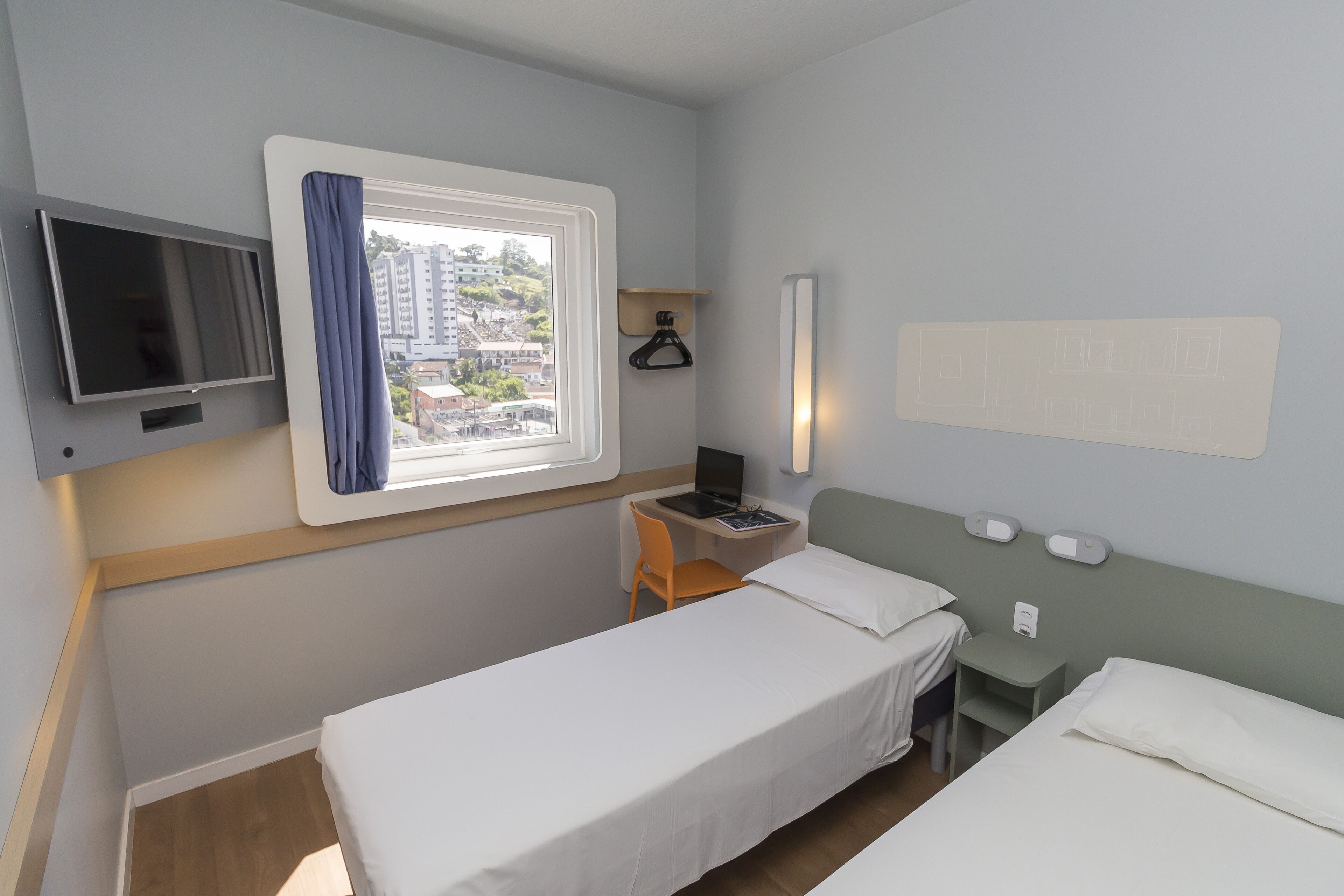 A hotel room with two single beds, a desk with a chair, a TV, a window with curtains, and a wall-mounted light.