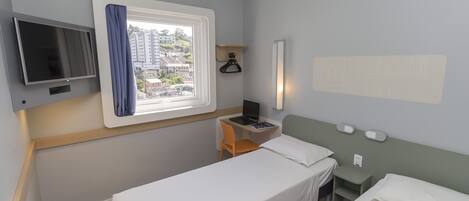 A hotel room with two single beds, a desk with a chair, a TV, a window with curtains, and a wall-mounted light.