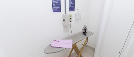 A small room with a foldable ironing board, an iron, and a pink towel. There are two instructional signs on the wall.