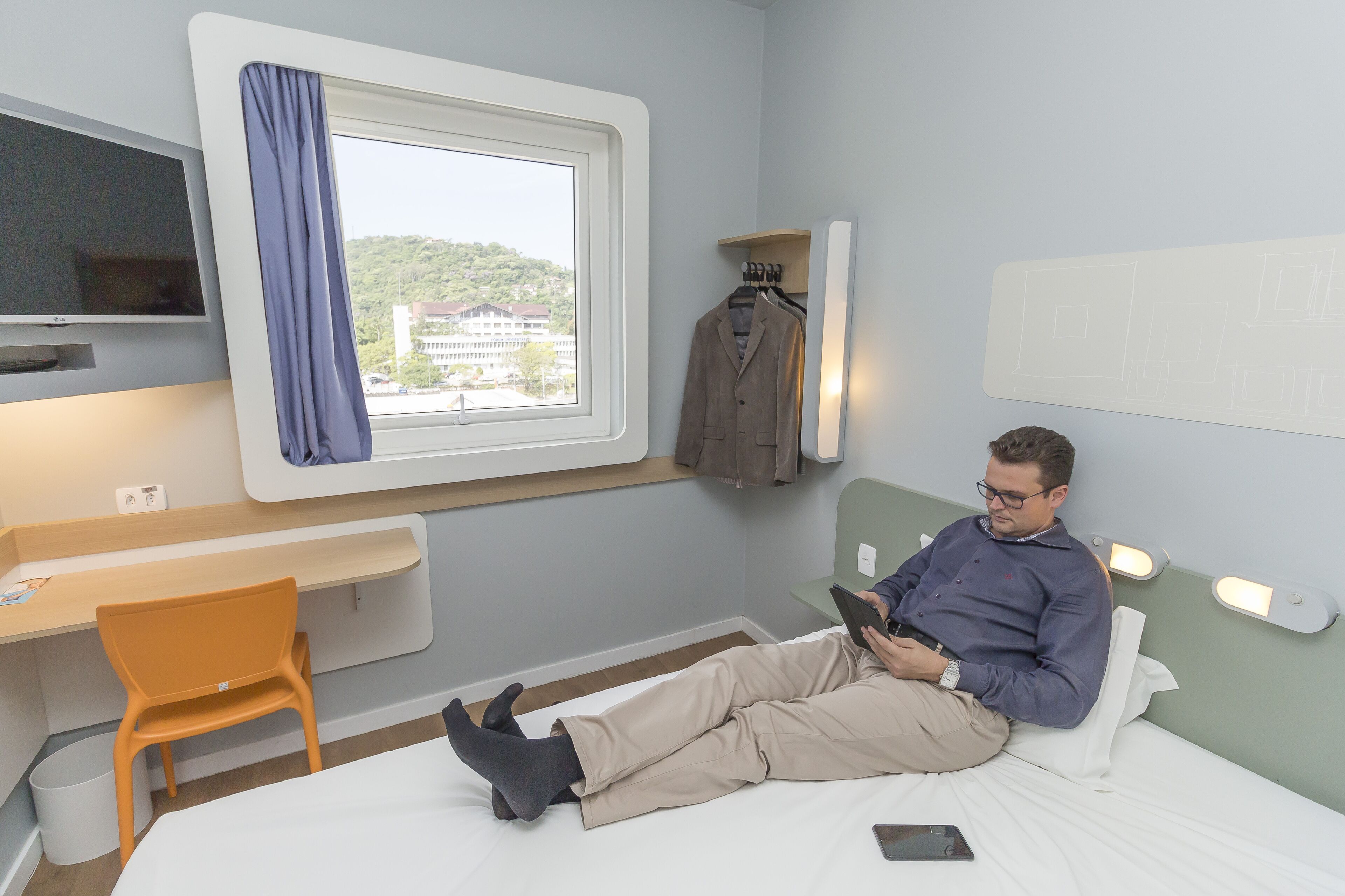 Quarto de hotel com cama, escrivaninha com cadeira, televisão, janela com vista e um cabideiro.