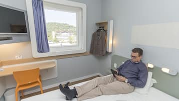 A hotel room with a bed, a desk with a chair, a TV, a window with a view, and a coat rack.