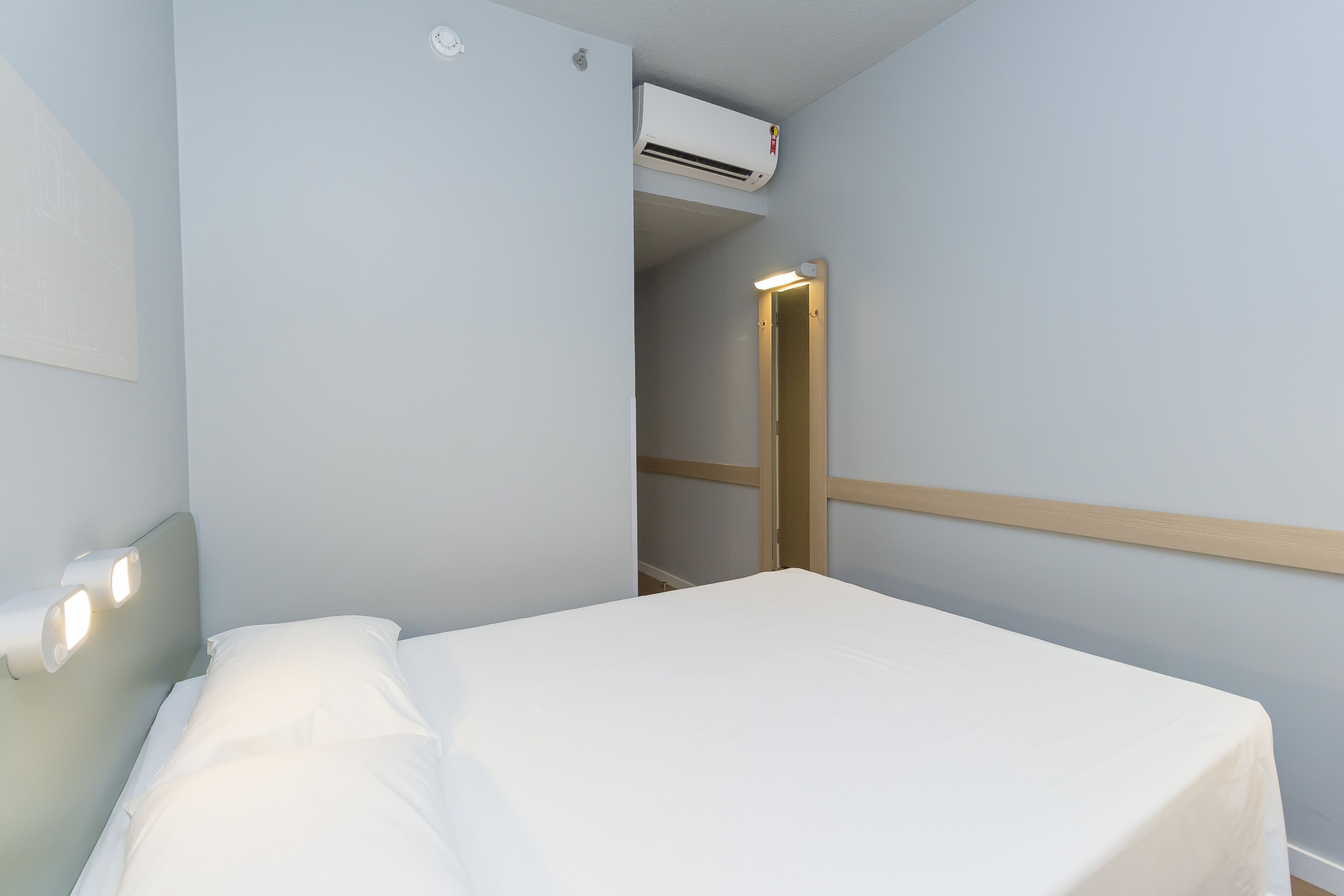 Una cama bien hecha en una habitación con paredes azul claro, cabecera de madera y unidad de aire acondicionado montada en la pared.