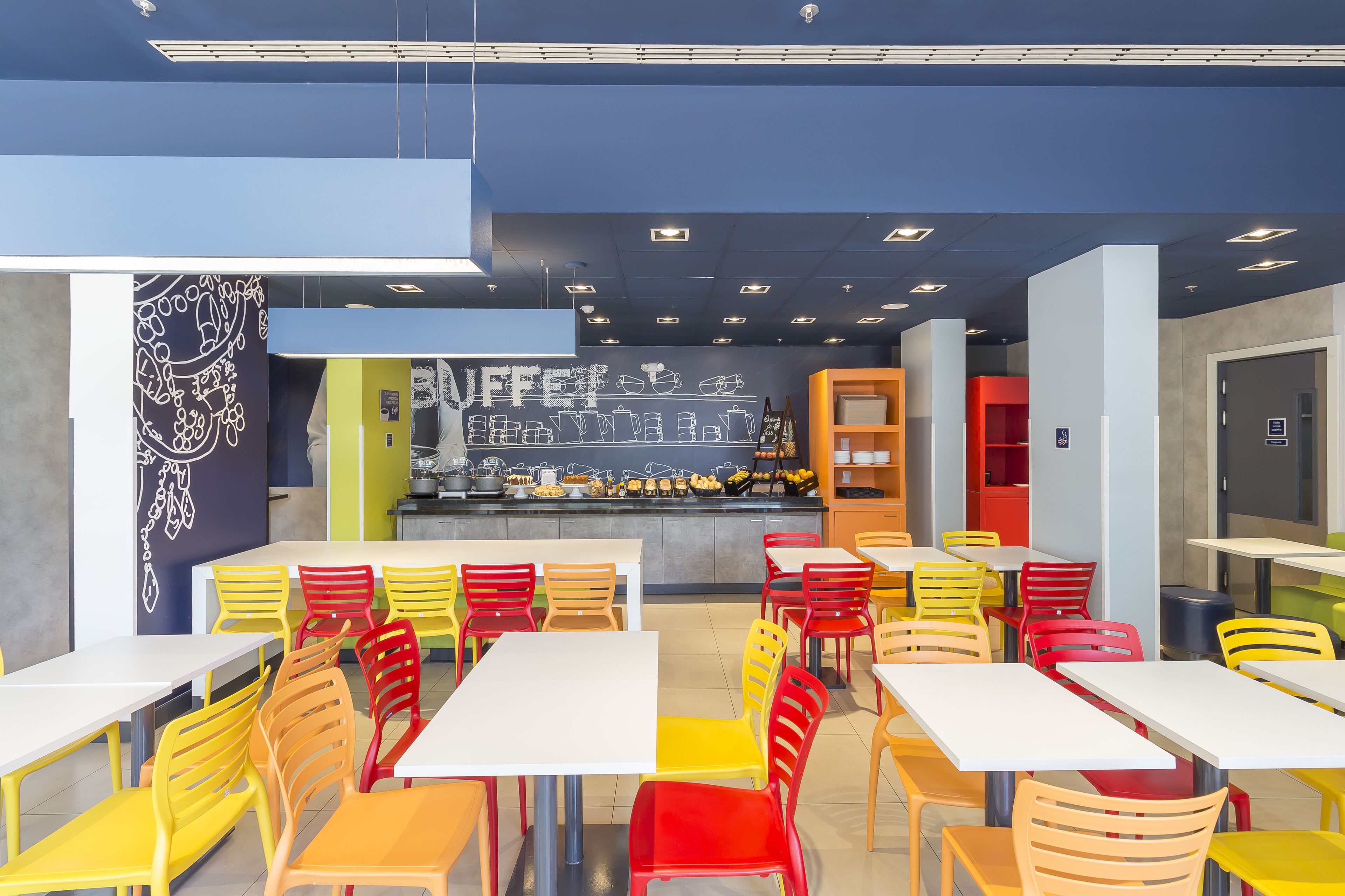 Eine moderne Cafeteria mit bunten Stühlen und Tischen, einer Kreidetafel mit Speisekarte und einem Küchenbereich im Hintergrund.