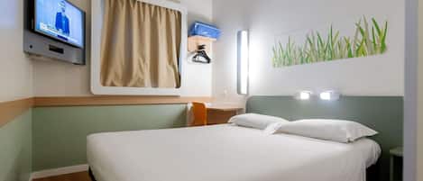 Una habitación de hotel con cama, televisión montada en la pared, ventana con cortinas y lámpara de pared.