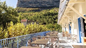 An outdoor terrace with tables and chairs, overlooking a forested hillside and a prominent cliff. -  Les Lavandes (Remuzat)