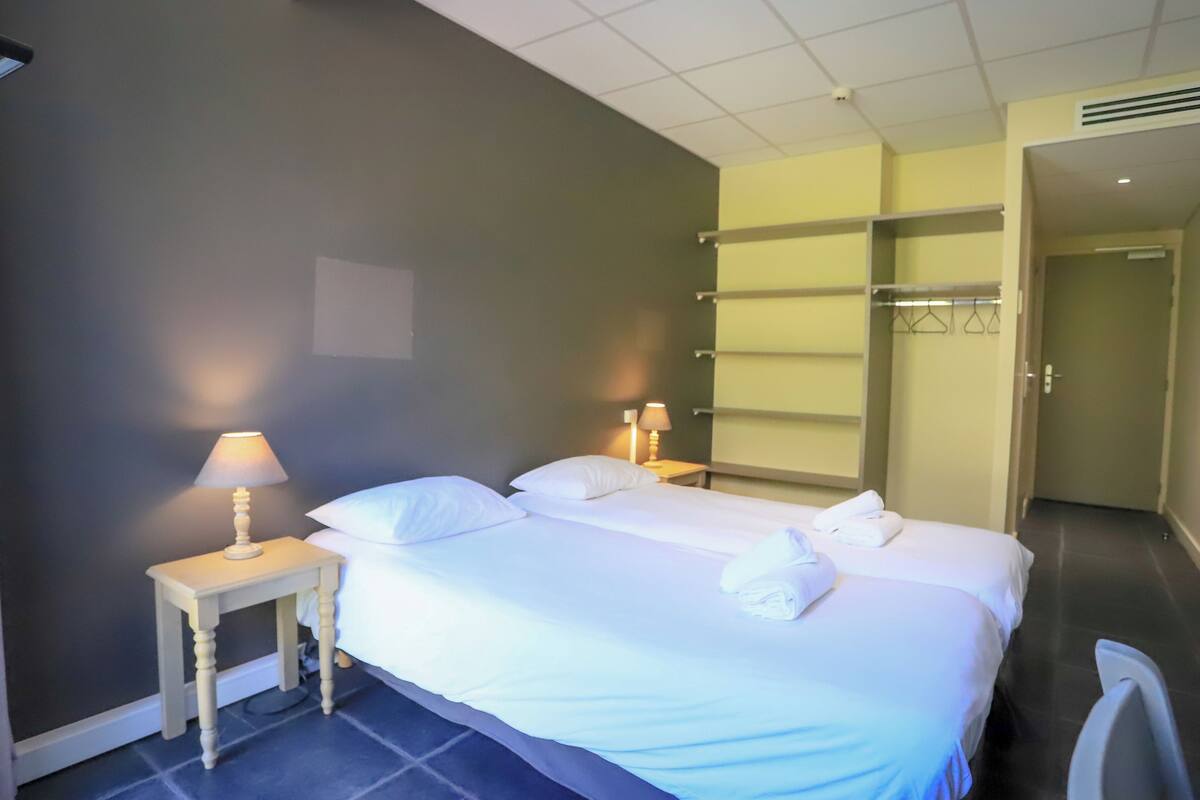 A hotel room with two beds, a small table with a lamp, a wardrobe, and a door leading to another room.