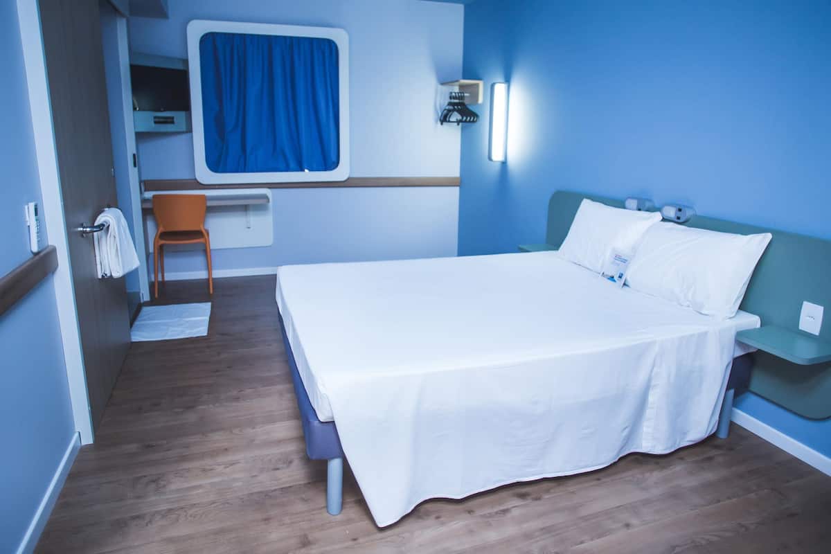 Quarto de hotel com cama, escrivaninha, cadeira, janela com cortinas e luminária na parede.