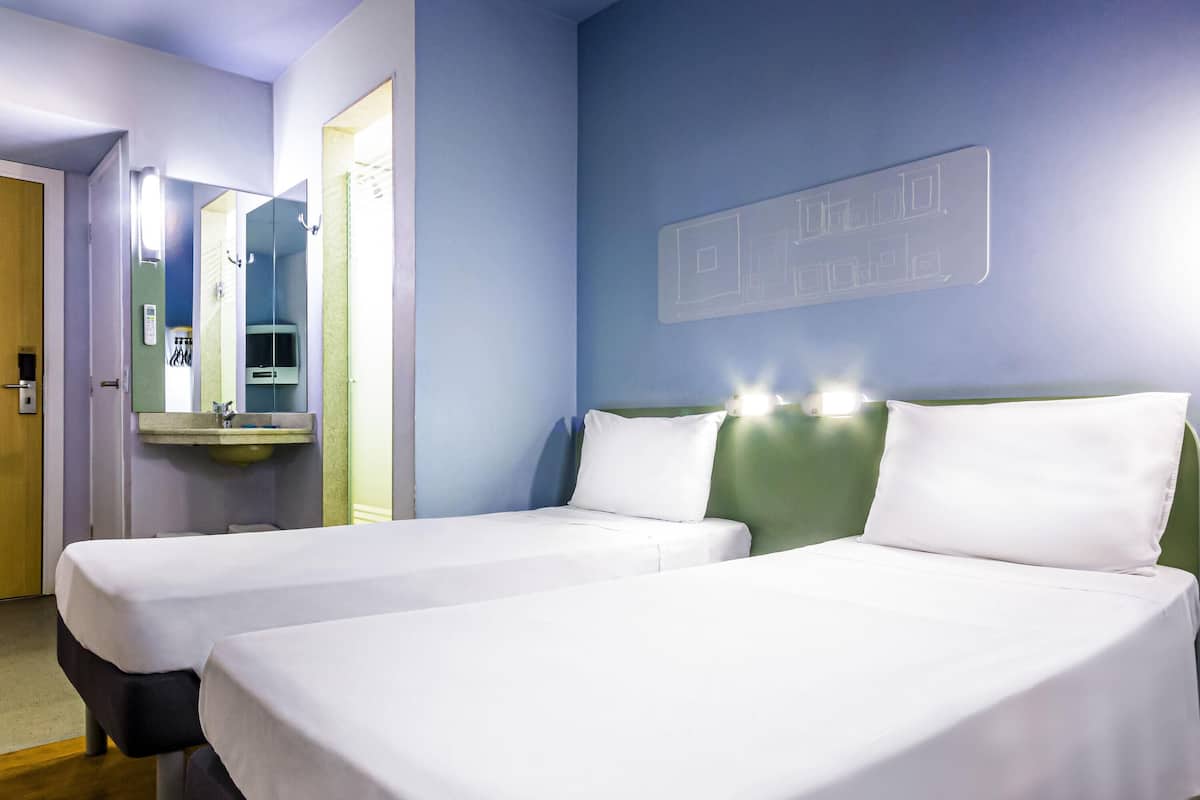 Quarto de hotel com duas camas, um espelho, uma pia e um candeeiro fixado na parede.