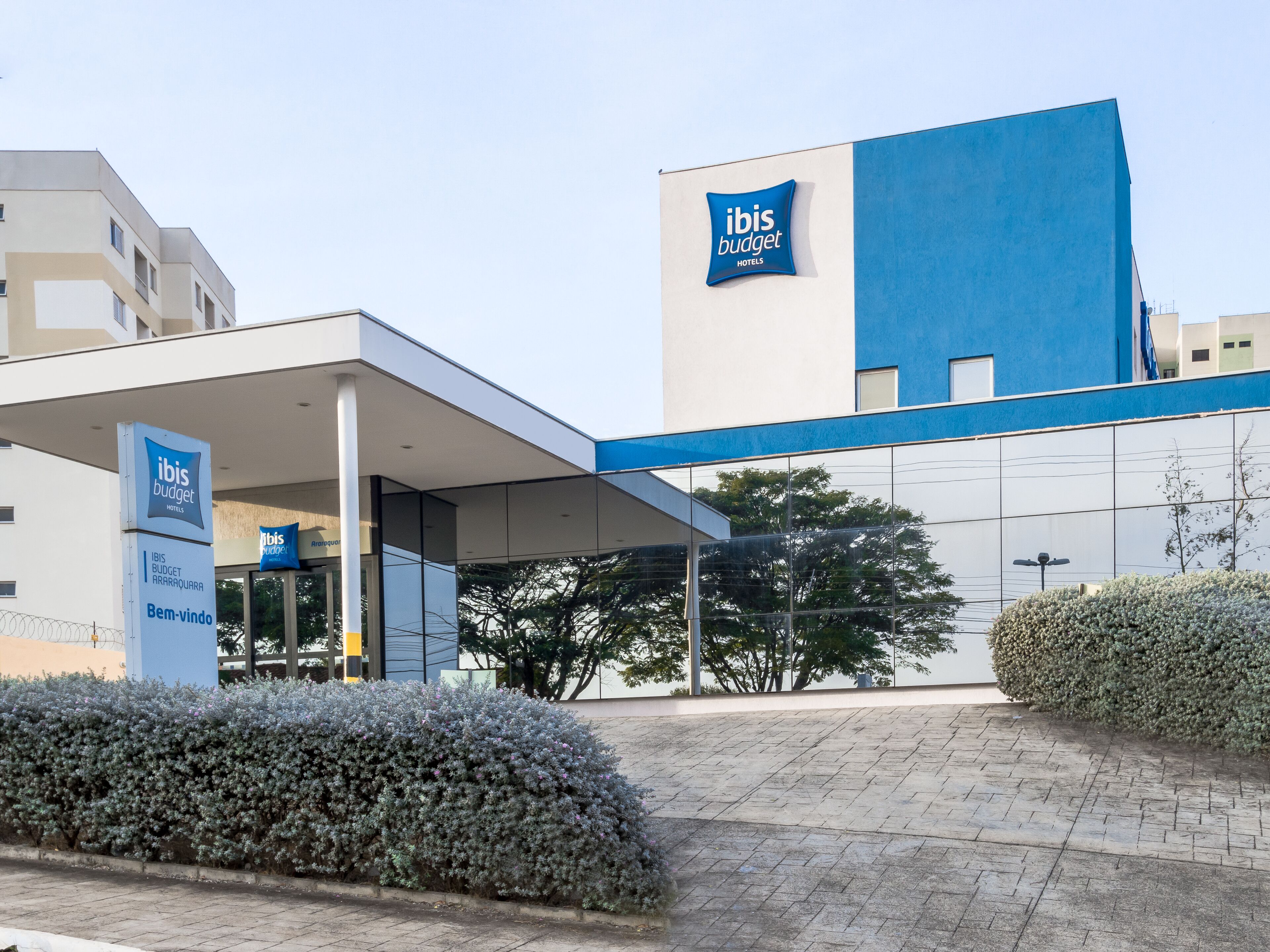 Una entrada de hotel moderna con fachada azul y blanca, un espacio exterior cubierto y un seto bien cuidado al frente.