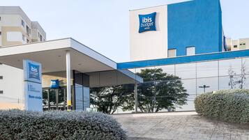 Una entrada de hotel moderna con fachada azul y blanca, un espacio exterior cubierto y un seto bien cuidado al frente.
