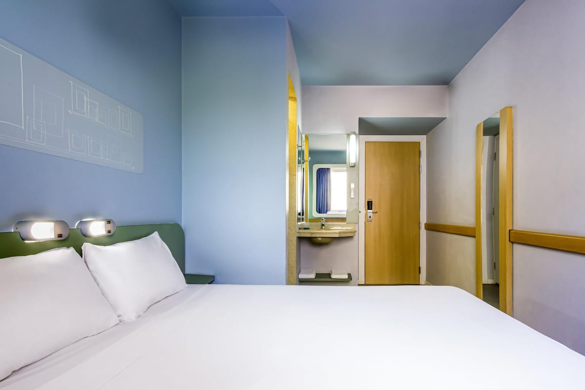Quarto de hotel com uma cama, banheiro com pia e espelho, e porta que dá para outro cômodo.