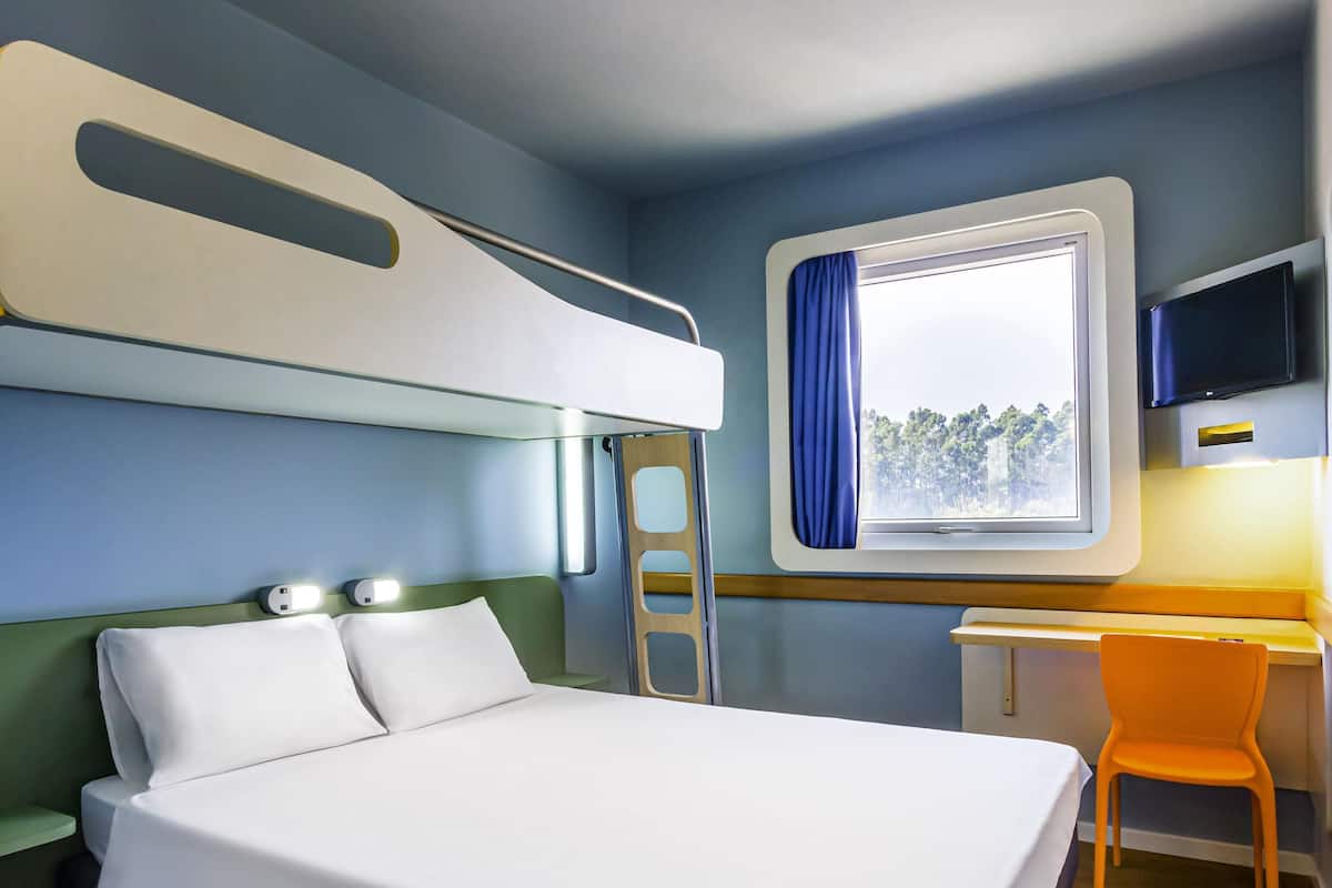 Uma cama beliche com escrivaninha e cadeira, uma televisão e uma janela com vista para árvores.