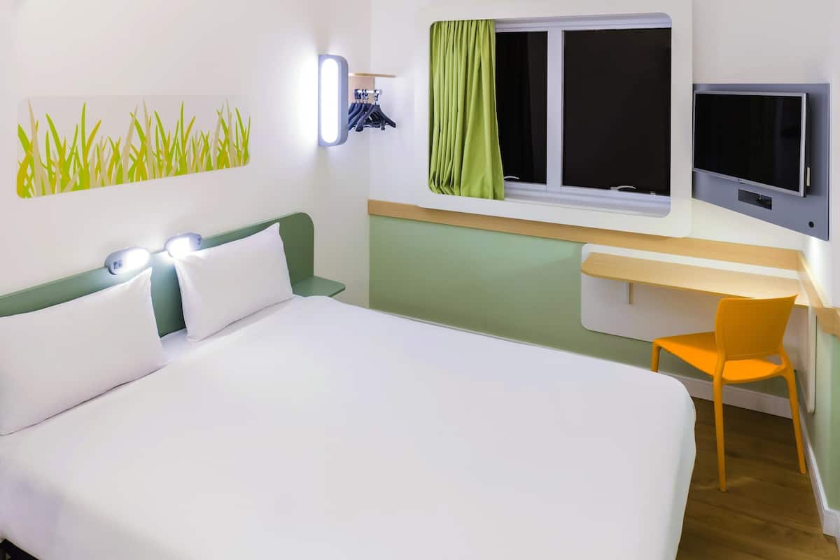 Habitación de hotel con cama, escritorio, silla, televisión y ventana con cortinas.