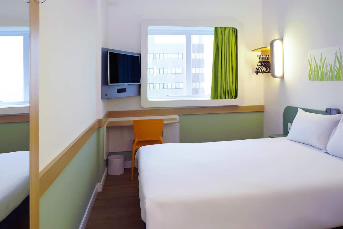 Hotellihuone, jossa on sänky, työpöytä tuolilla, televisio, ikkuna verhoilla ja seinälle asennettu valaisin.