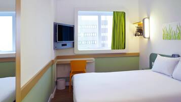 Habitación de hotel con cama, escritorio con silla, televisor, ventana con cortinas y lámpara fijada a la pared.