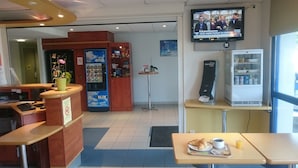 Un restaurant équipé d’un téléviseur diffusant les actualités, d’un distributeur automatique, d’un distributeur de jeux « Just One » et d’une table où l’on sert le petit-déjeuner, avec des croissants et du jus de fruits.