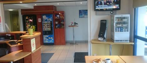 Un restaurante con una televisión mostrando noticias, una máquina expendedora, un dispensador Just One y una mesa de desayuno con croissants y jugo.