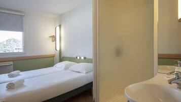 A hotel room with two single beds, a window, a bathroom with a sink and mirror, and a view of the outdoors.