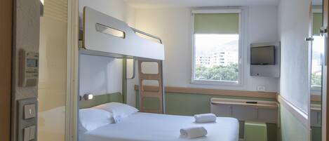 Habitación de hotel con literas, una cama individual, un baño con inodoro, lavabo, espejo, ventana con vistas a edificios y un televisor en la pared.