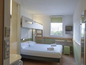 A hotel room with a bunk bed, a single bed, a bathroom with a toilet, a sink, a mirror, a window with a view of buildings, and a TV on the wall. - ibis budget Ajaccio (Ajaccio)