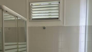 Ein Badezimmer mit Duschkabine aus Glas, einem Handtuchhalter mit Handtuch, einem Fenster mit Jalousien und einem Waschbecken mit Spiegel.