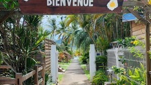 Un sentier mène à une arche en bois sur laquelle est affiché un panneau indiquant « BIENVENUE », le tout étant entouré d’une végétation luxuriante et de plantes tropicales.
