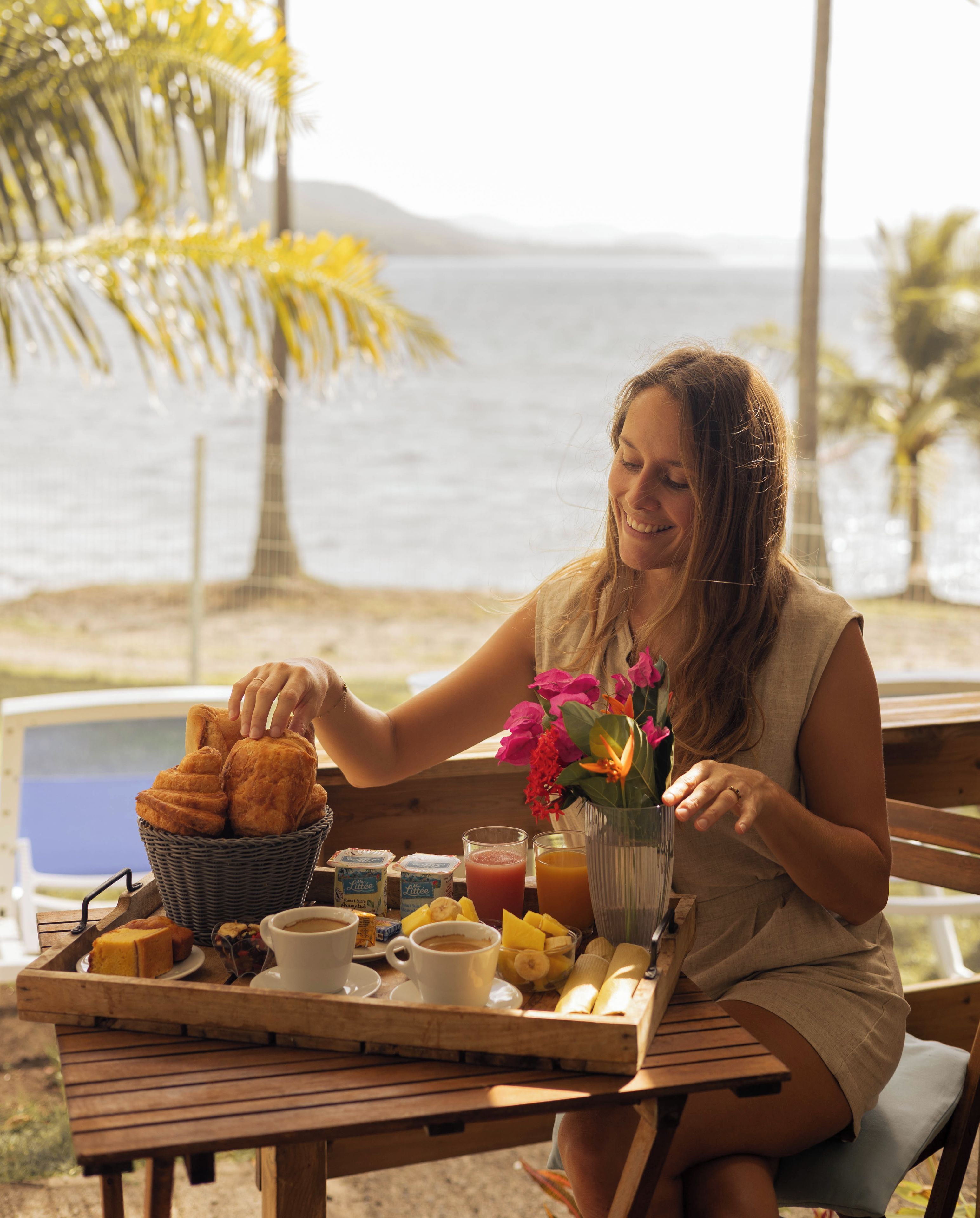 A person seated at a wooden table with a tray of breakfast items, including bread, fruit, and beverages, overlooking a body of water.