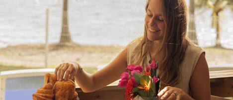 A person seated at a wooden table with a tray of breakfast items, including bread, fruit, and beverages, overlooking a body of water.