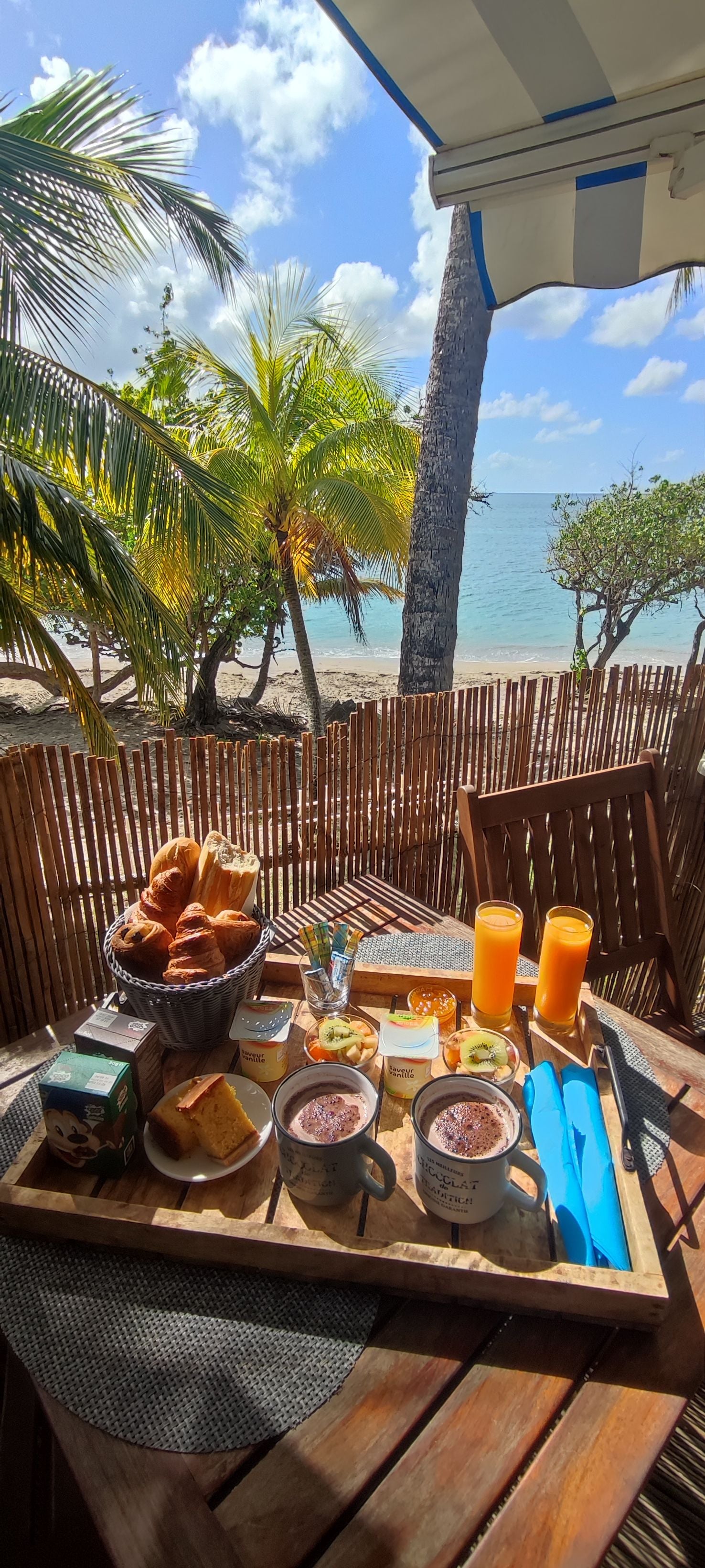 A wooden table with breakfast items, including bread, fruit, and drinks, set against a backdrop of a beach and palm trees.