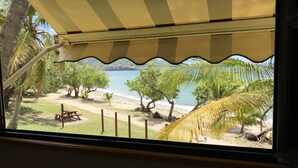 Une vue sur la plage, prise depuis une fenêtre ornée d’un auvent à rayures, avec des palmiers et une zone sablonneuse où se trouve un banc.