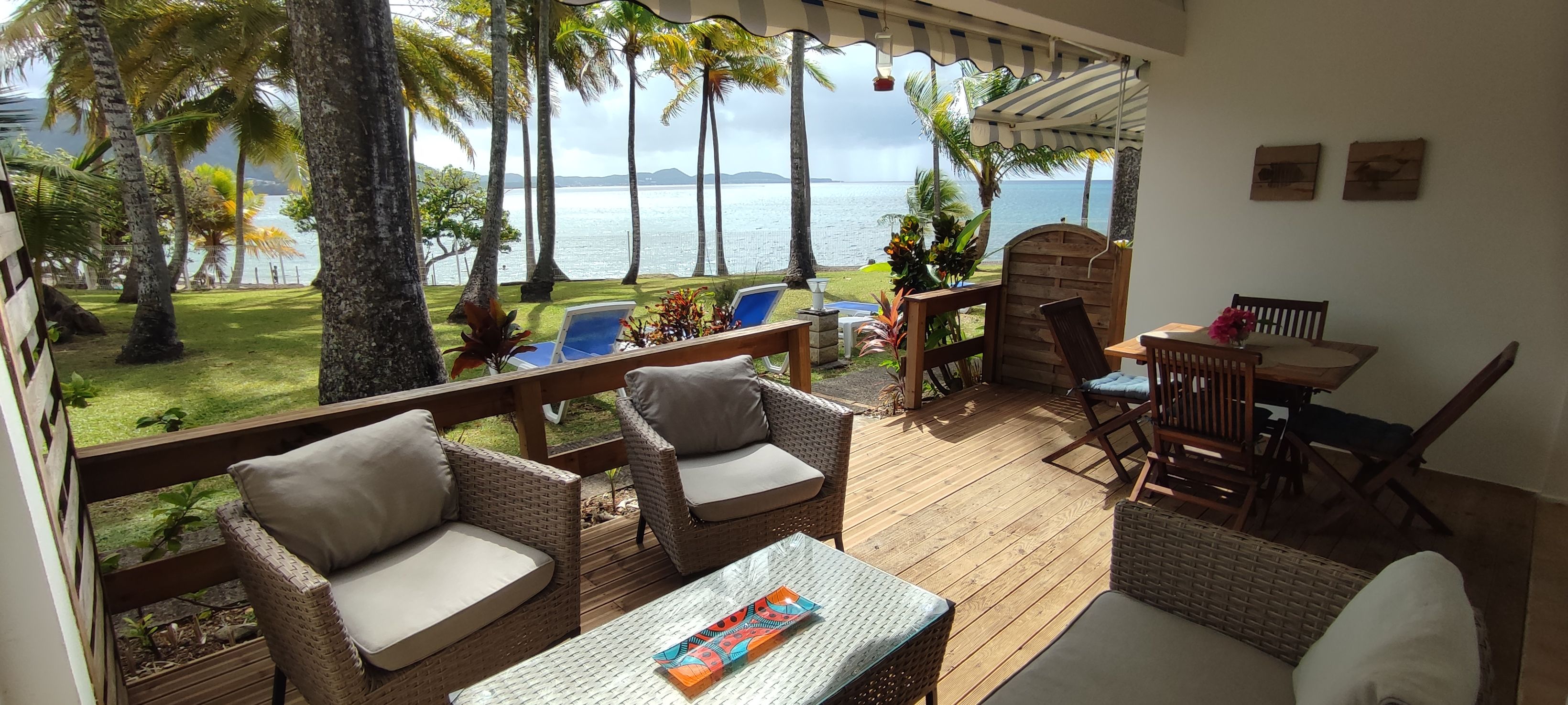 A covered outdoor seating area with wicker furniture, a table, and chairs, overlooking a body of water with palm trees.