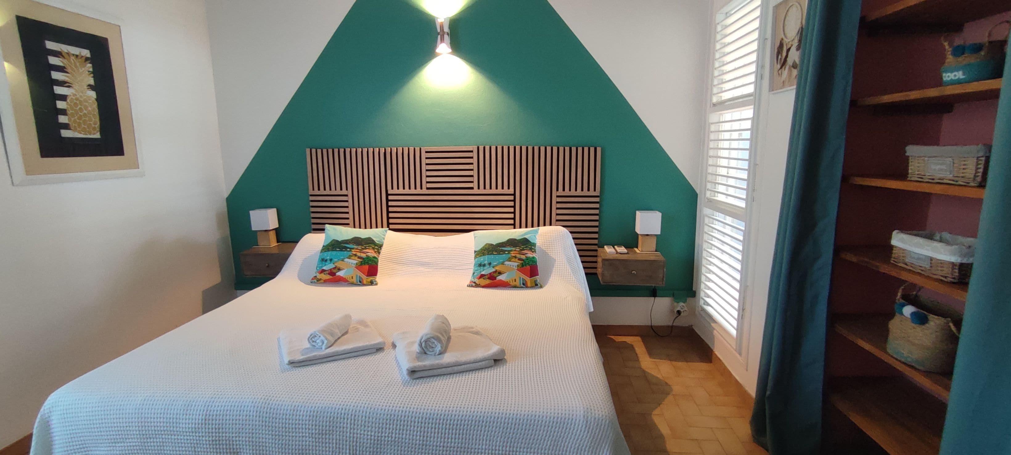 A hotel room with a bed, two pillows, a nightstand, a lamp, and a shelf with storage boxes.