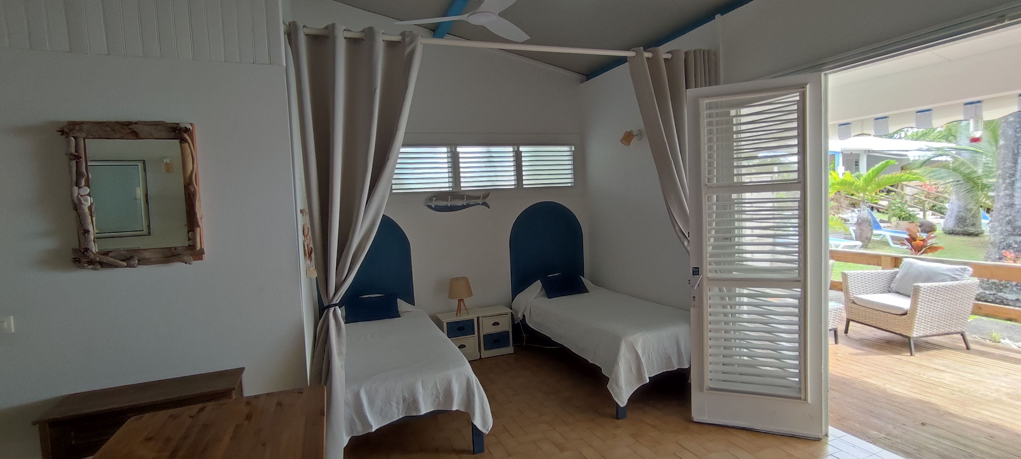 A room with two beds, a ceiling fan, a window with blinds, and a view of an outdoor area with furniture.