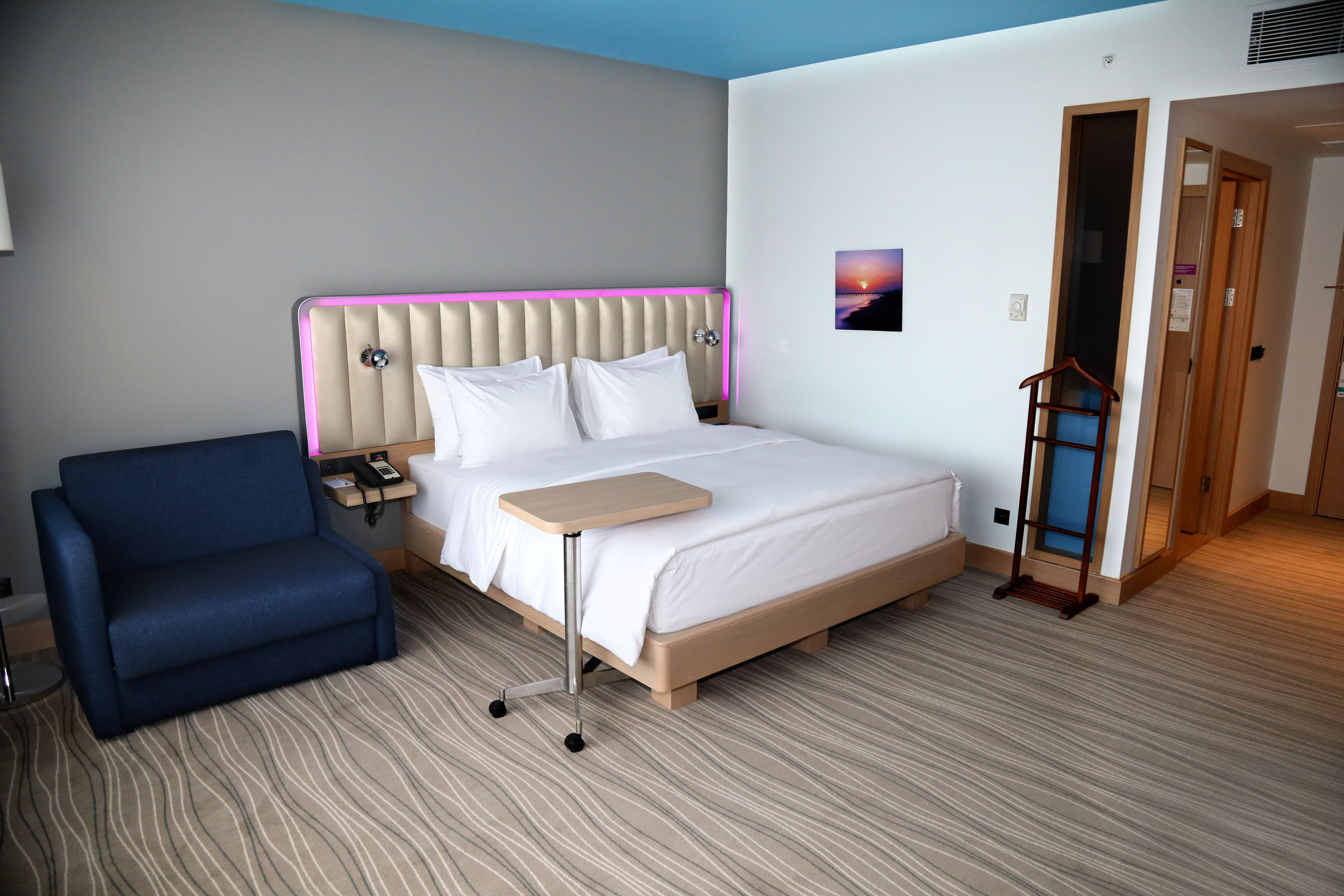A hotel room with a bed, a blue armchair, a bedside table, and a picture on the wall.