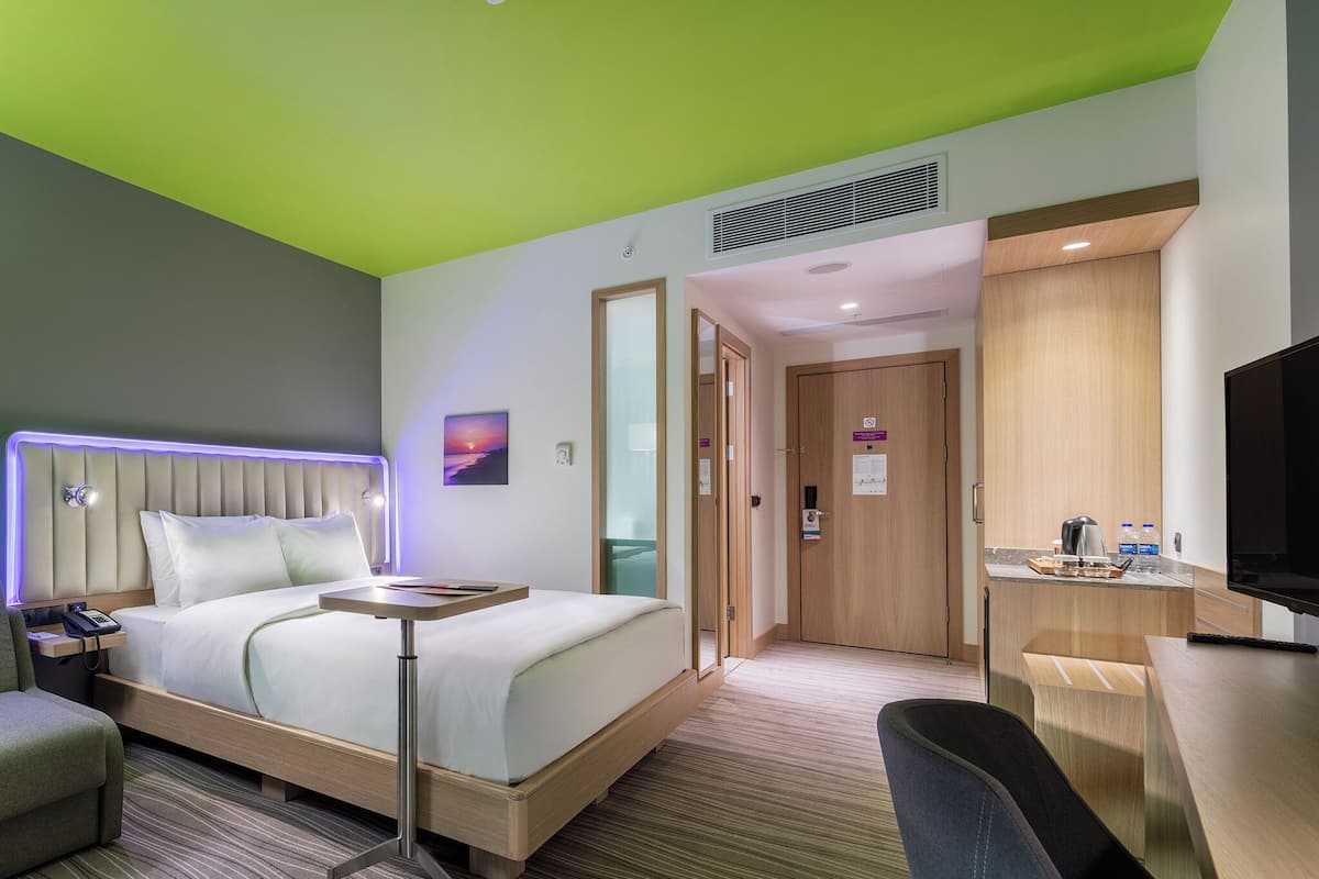 A hotel room with a bed, bedside table, chair, desk, TV, and a bathroom with a shower and toilet.